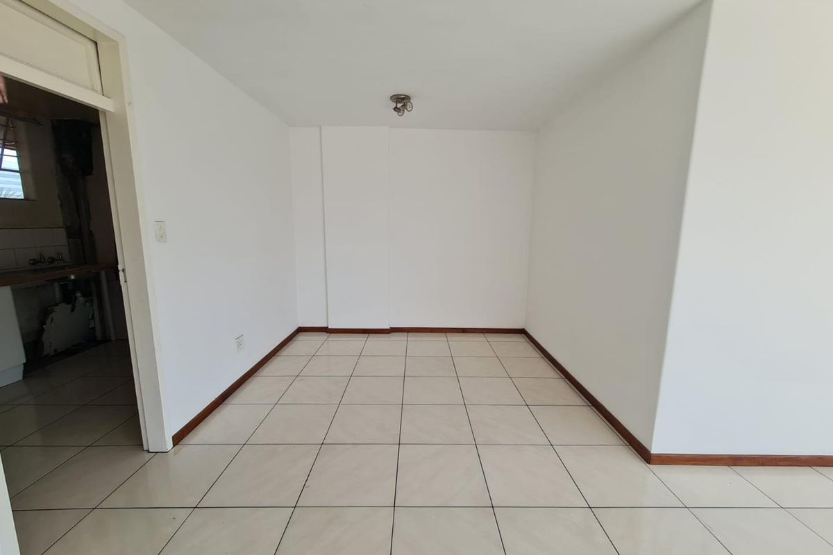 An empty, neutrally finished living area featuring light-colored floor tiles and white walls, with an adjacent view into a secondary utility or kitchen space.