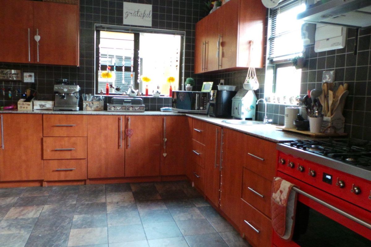 A functional kitchen featuring wooden cabinetry, dark tiled splashbacks, tiled flooring, and a prominent red freestanding gas stove with an extractor fan.