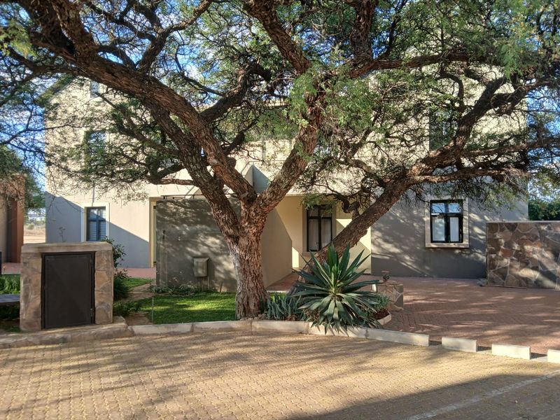 Front elevation of the apartment building featuring a paved driveway, stone cladding accents, and a large mature tree providing shade to the entrance area.
