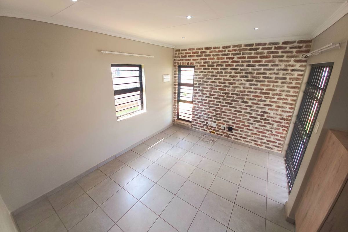 A bright, empty living room featuring neutral floor tiles, recessed ceiling lighting, and a distinctive exposed brick feature wall.