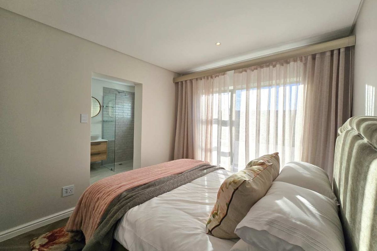 A well-maintained bedroom featuring neutral tones, floor-to-ceiling curtains, and an en-suite bathroom with a glass-enclosed shower and modern vanity.