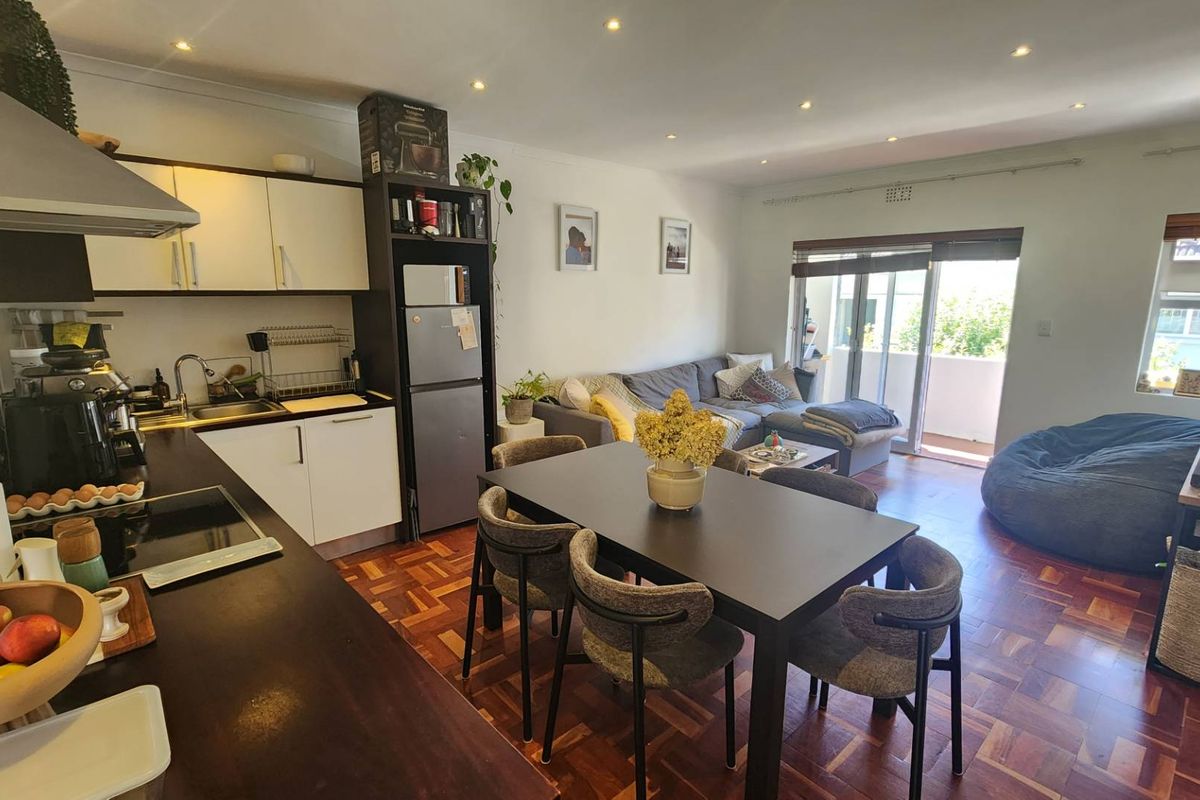 Open-plan living area featuring warm parquet flooring, recessed lighting, and a functional kitchen with modern cabinetry, flowing seamlessly to a private balcony.