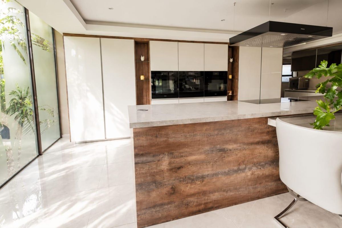 Modern kitchen featuring a large central island with stone countertop, integrated ovens, sleek cabinetry, and floor-to-ceiling glass walls overlooking a private garden.