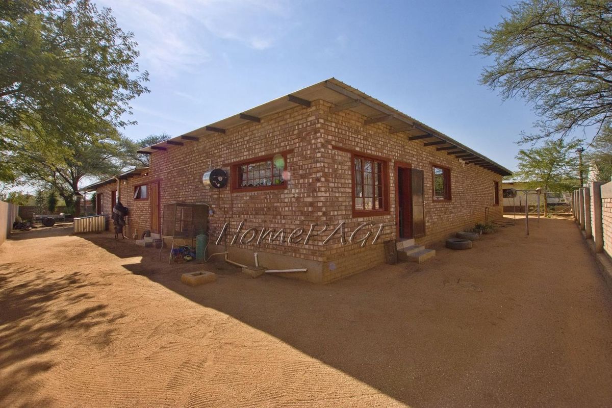 Exterior view of a single-story face-brick house featuring a simple roof structure and basic window frames, set on a sandy plot.