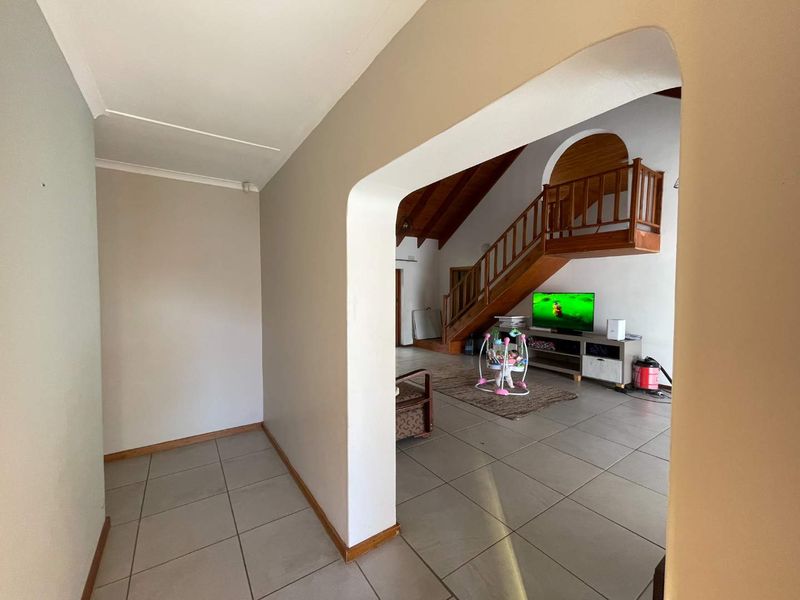 Open plan living area featuring tiled flooring, a prominent wooden staircase with balustrade, and a vaulted ceiling, viewed through an arched entryway.