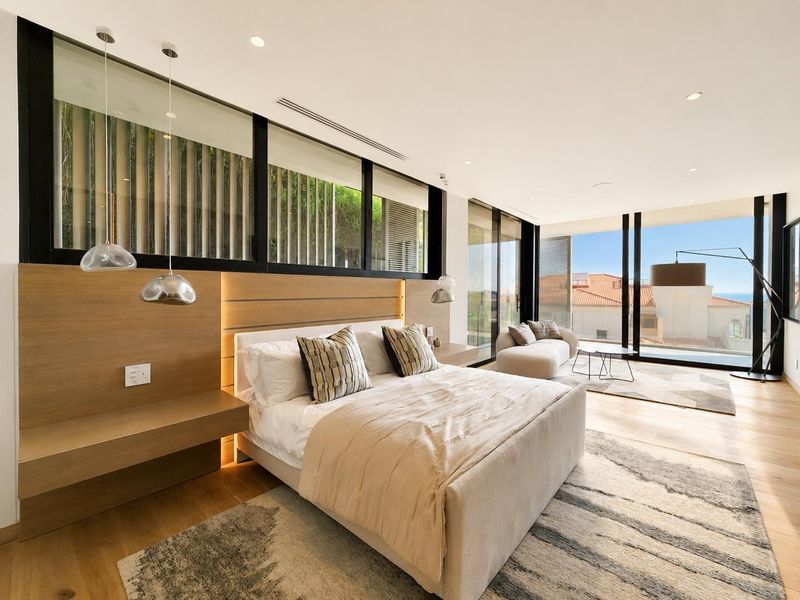 Spacious modern bedroom featuring timber flooring, built-in headboard with integrated lighting, and floor-to-ceiling glass doors opening to a scenic view.