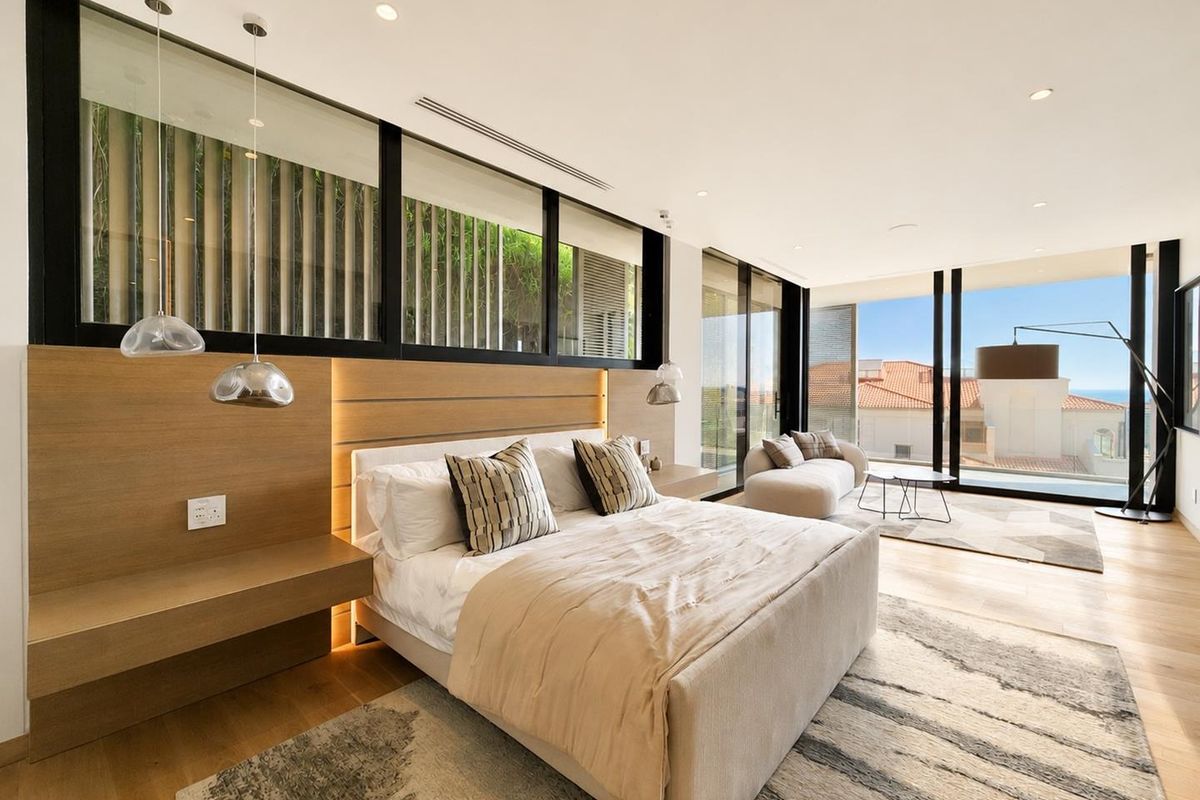 Spacious modern bedroom featuring timber flooring, built-in headboard with integrated lighting, and floor-to-ceiling glass doors opening to a scenic view.