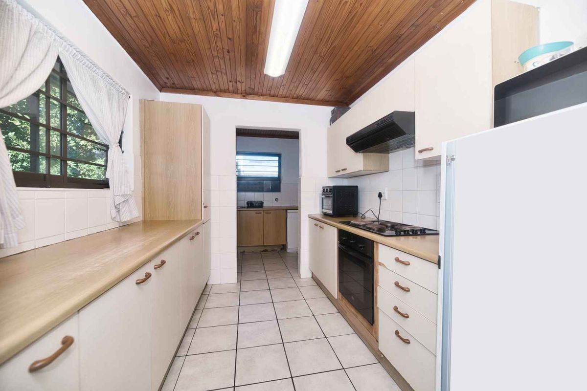 A functional kitchen featuring built-in cabinetry, a hob, oven, and extractor fan, with a tiled floor and timber ceiling, leading to a secondary prep area.