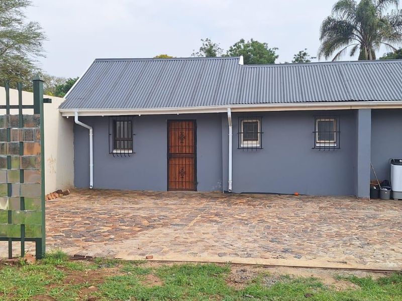 Front exterior view of a cottage featuring a corrugated metal roof, paved driveway, and secure perimeter fencing with security bars on windows.