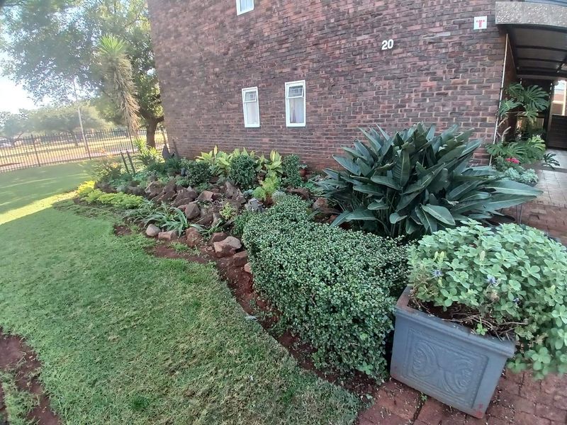 A well-maintained front garden area featuring established shrubs, rock landscaping, and a brick building facade with a paved walkway.