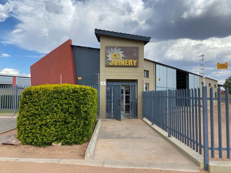 Front entrance of an industrial property featuring secure steel palisade fencing, electric security measures, and a paved walkway leading to the main building entrance.