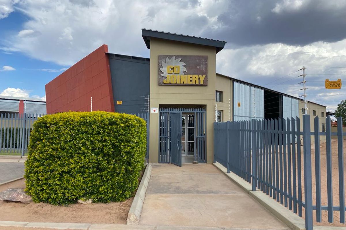 Front entrance of an industrial property featuring secure steel palisade fencing, electric security measures, and a paved walkway leading to the main building entrance.