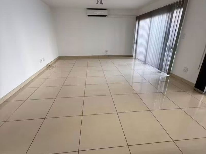 A bright, empty living room featuring large format floor tiles, a wall-mounted air conditioning unit, and sliding glass doors with vertical blinds.