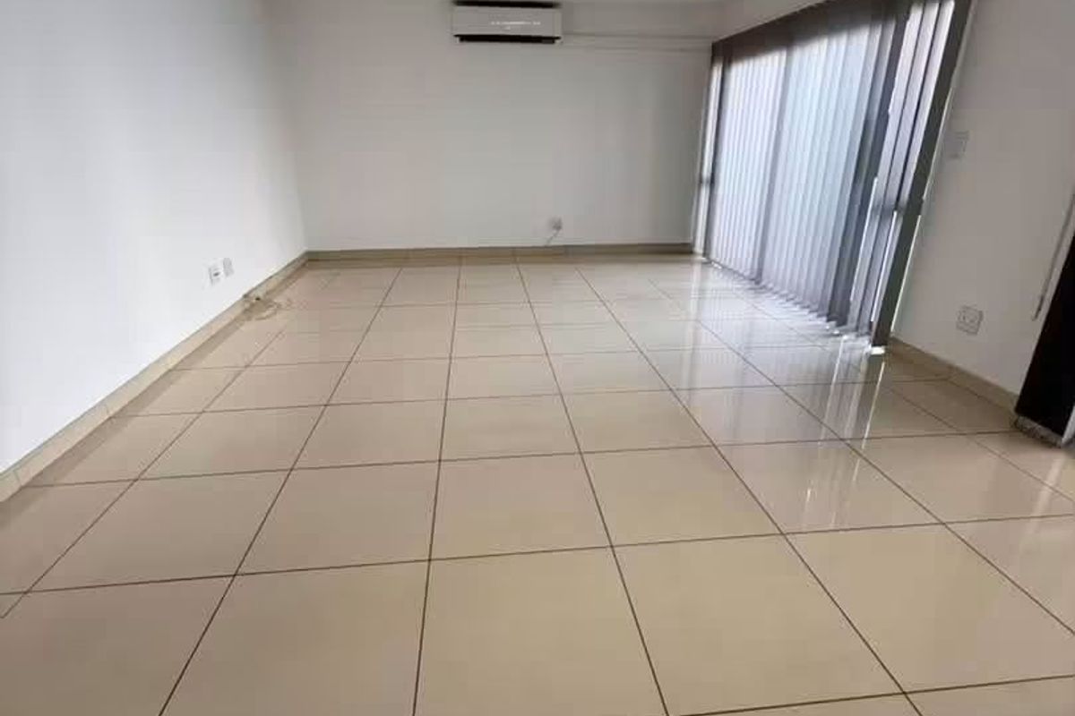 A bright, empty living room featuring large format floor tiles, a wall-mounted air conditioning unit, and sliding glass doors with vertical blinds.