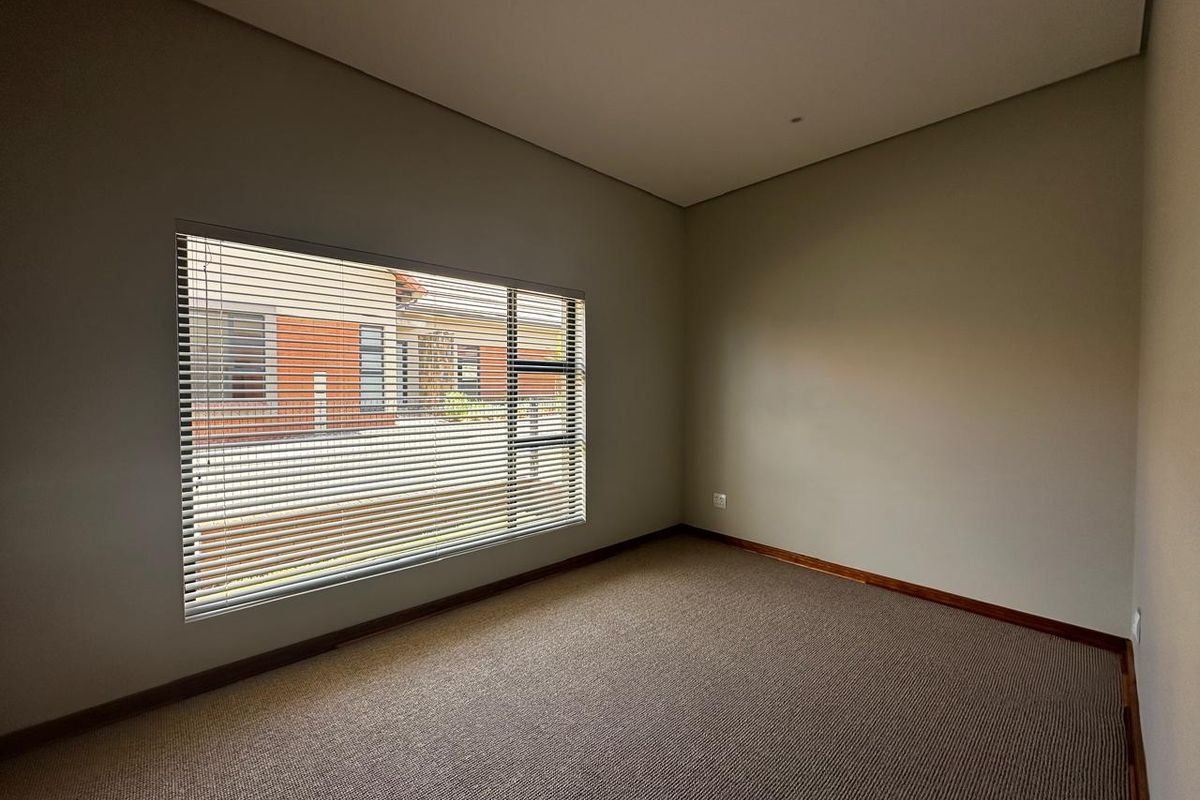 A bright, empty bedroom featuring neutral wall tones, fitted carpeting, and a large window with installed horizontal blinds.