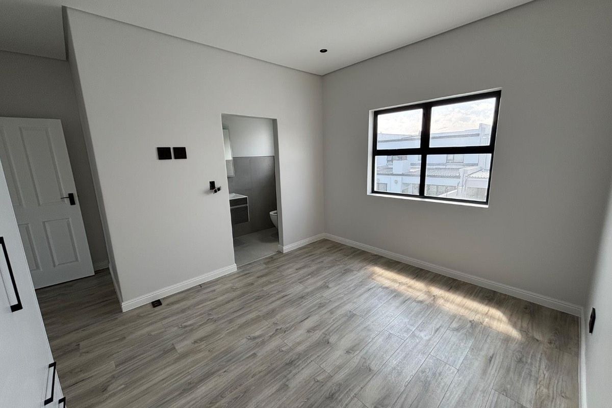 A bright, modern, empty bedroom featuring light wood-look laminate flooring, neutral walls, and an en-suite bathroom entrance.