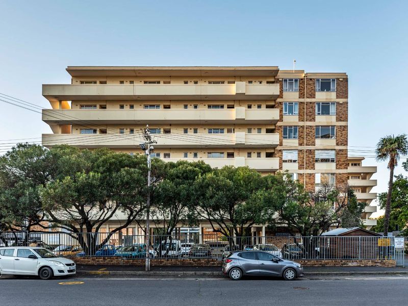 Front elevation of a multi-story apartment building featuring tiered balconies, brick facade detailing, and secure off-street parking behind metal fencing.