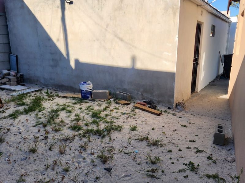 Rear exterior area featuring an unfinished concrete wall, security gate, and sandy ground requiring landscaping and surface finishing.