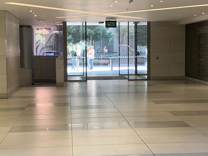 A spacious and modern office building entrance lobby featuring polished tiled flooring, recessed ceiling lighting, and large glass sliding doors.