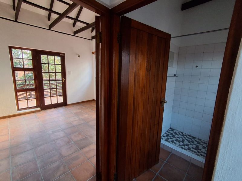 An empty bedroom featuring exposed timber ceiling beams, terracotta-style floor tiles, and an en-suite shower room with direct access to a balcony.