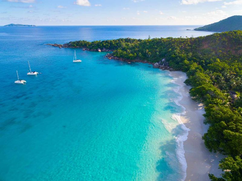 An aerial view of a pristine tropical coastline featuring white sandy beaches, lush vegetation, and turquoise ocean waters with anchored boats.