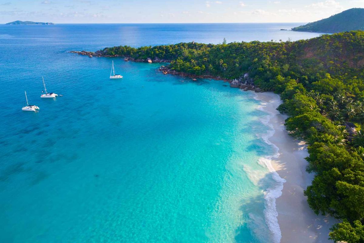 An aerial view of a pristine tropical coastline featuring white sandy beaches, lush vegetation, and turquoise ocean waters with anchored boats.