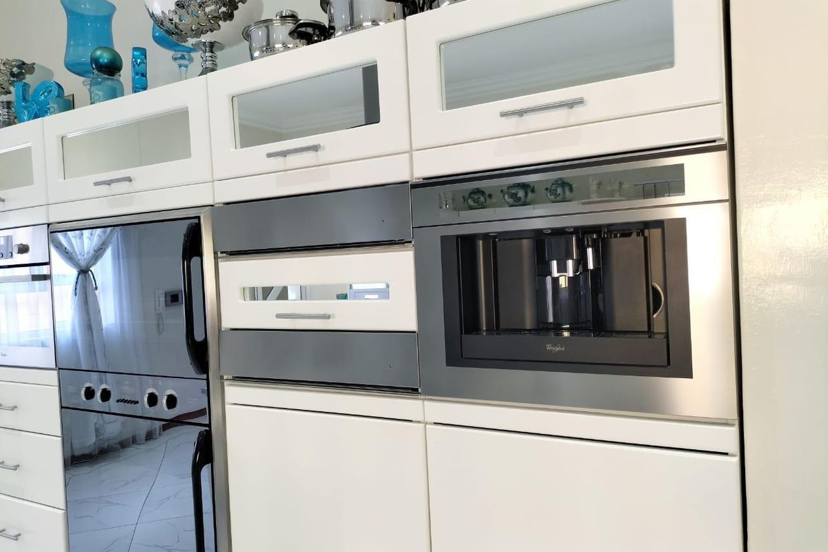 Modern kitchen cabinetry featuring integrated stainless steel appliances, including a built-in coffee machine and oven, with sleek white finishes and glass-fronted upper cabinets.