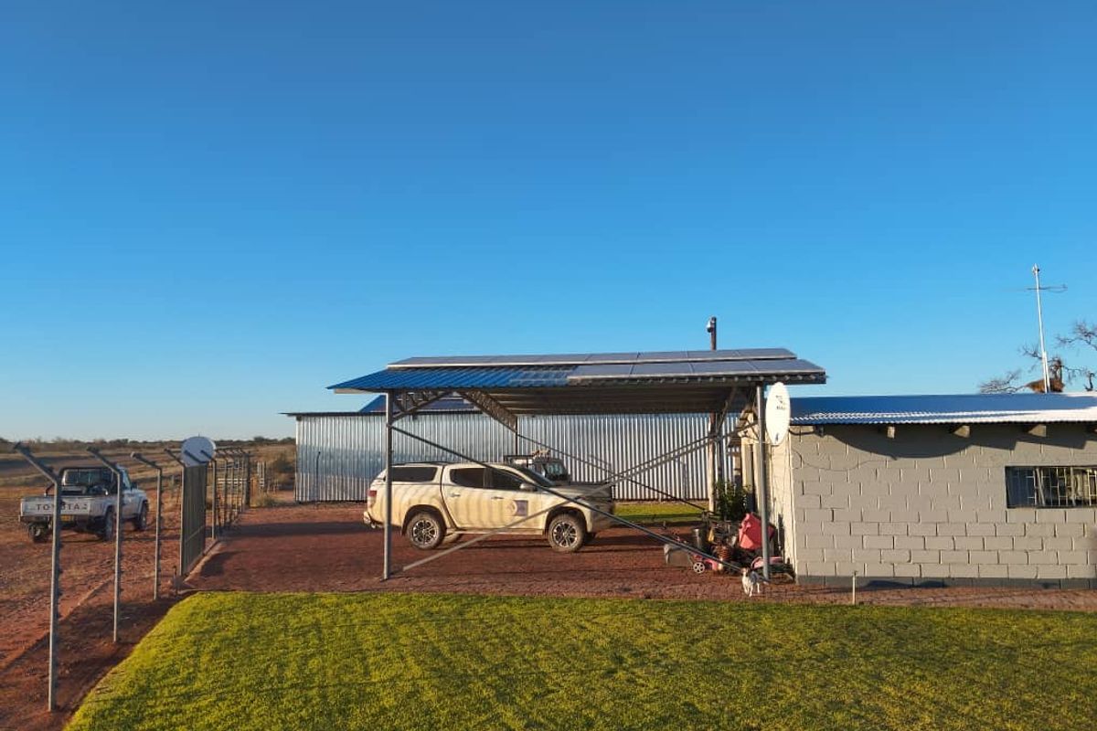 Exterior view of a small holding featuring a metal carport structure with integrated solar panels, adjacent to a concrete block building and fenced yard.