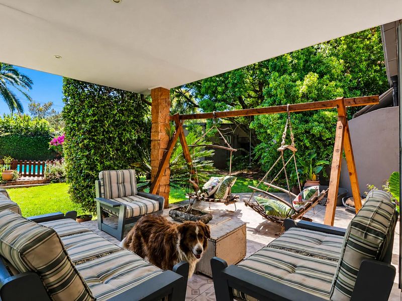 A covered outdoor patio area featuring stone-clad pillars and paved flooring, overlooking a lush garden with a glimpse of a swimming pool.
