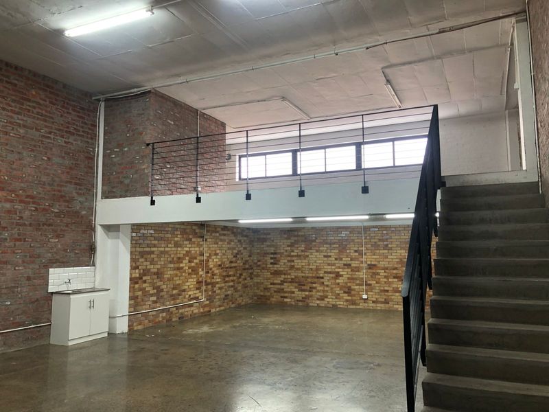 Spacious industrial-style open plan area featuring exposed brick walls, a mezzanine level with metal staircase, and a small kitchenette unit.
