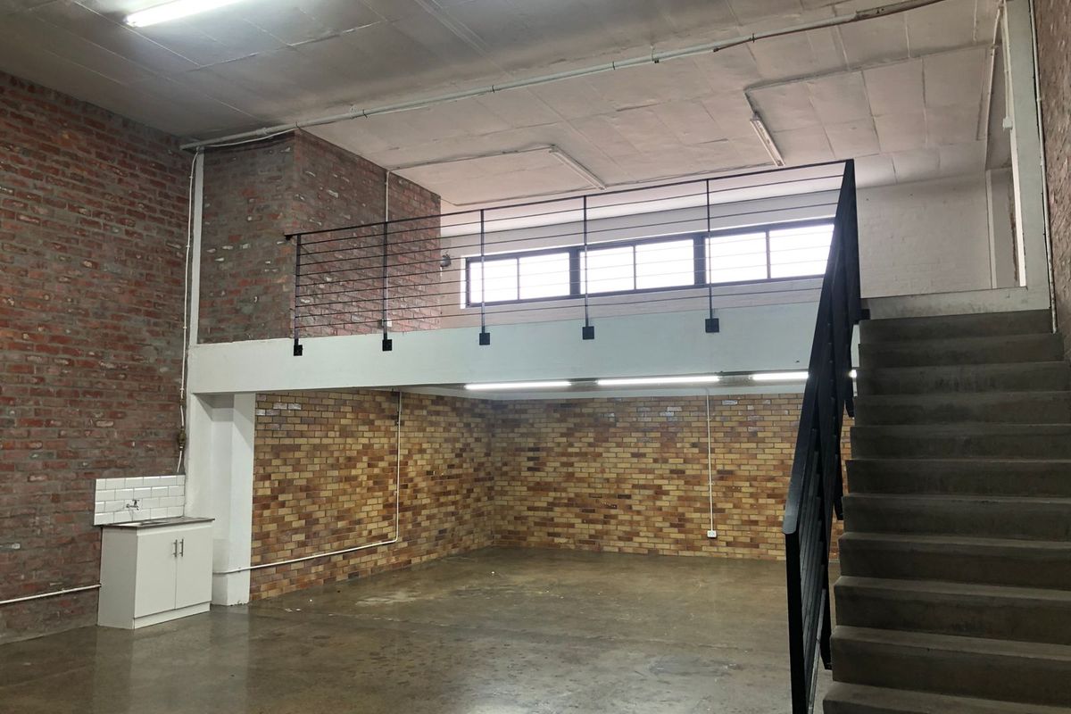 Spacious industrial-style open plan area featuring exposed brick walls, a mezzanine level with metal staircase, and a small kitchenette unit.