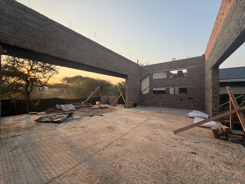 An unfinished open-plan living area under construction, featuring exposed brick walls, steel beams, and a concrete floor with views of the surrounding landscape.