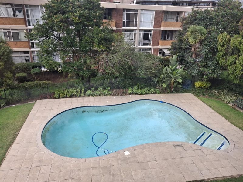 An elevated view of a communal swimming pool area featuring paved surrounds and established garden landscaping within an apartment complex.