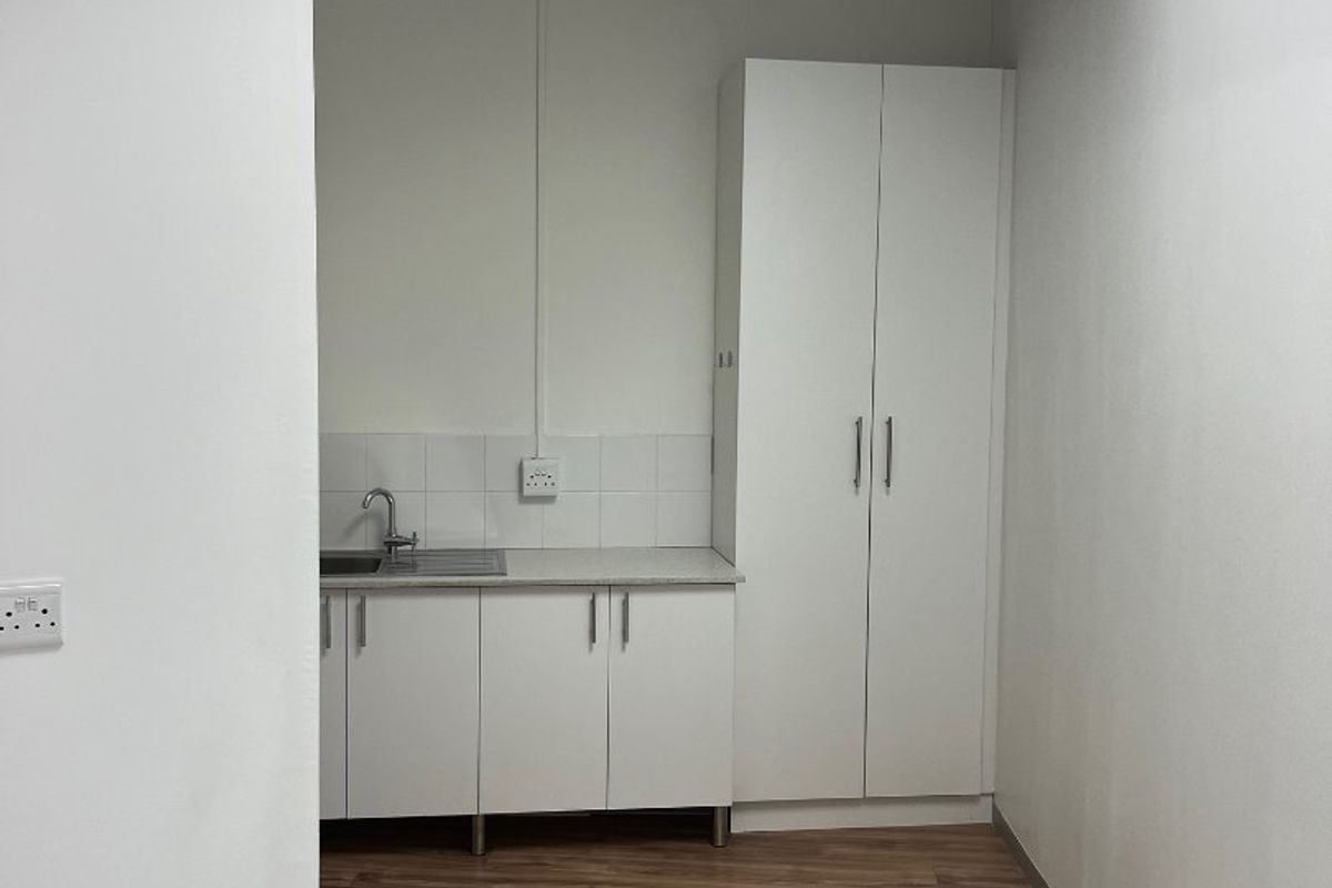 A clean, compact kitchenette area featuring white built-in cabinetry, a stainless steel sink, and wood-look flooring, suitable for office or retail utility use.