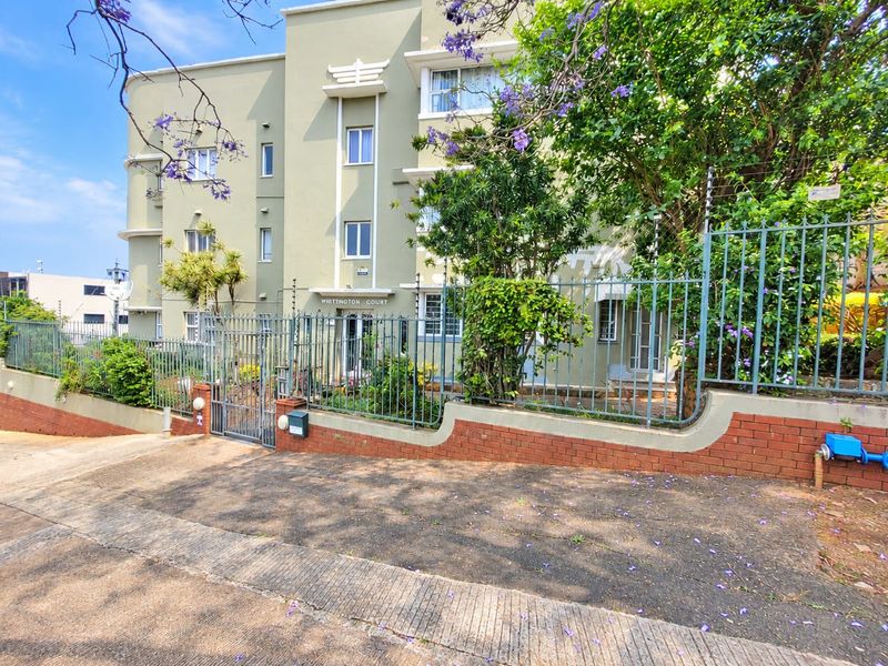 Front exterior view of Whittington Court apartment building featuring a secure gated entrance, brick boundary wall, and established landscaping.