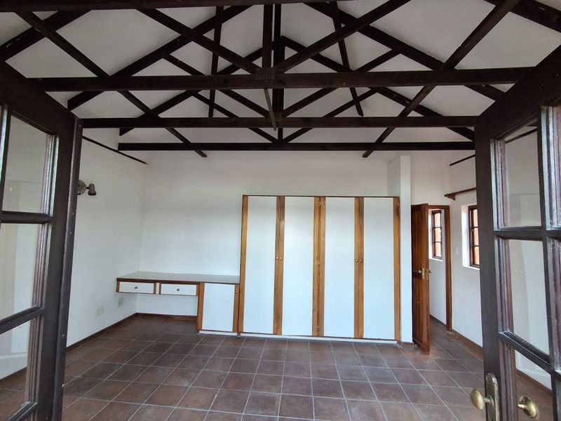 Spacious bedroom featuring exposed timber roof trusses, tiled flooring, built-in cupboards, and a built-in desk, accessible via double french doors.