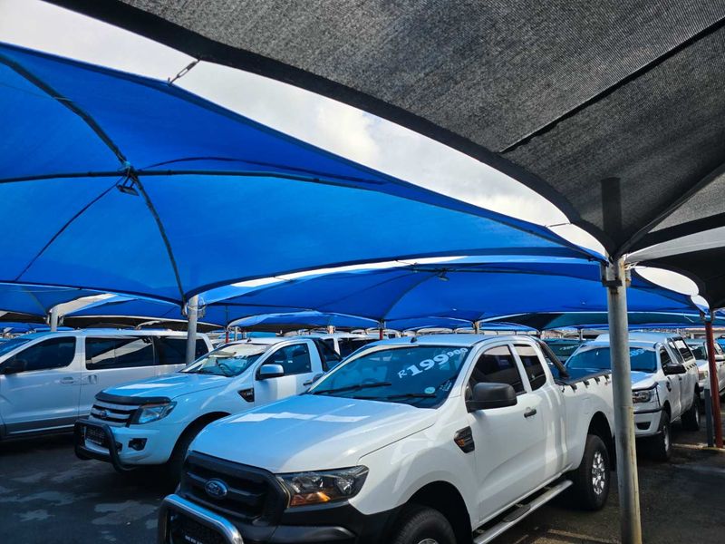 Outdoor parking area featuring a large blue shade cloth structure supported by steel poles, providing cover for multiple vehicles.