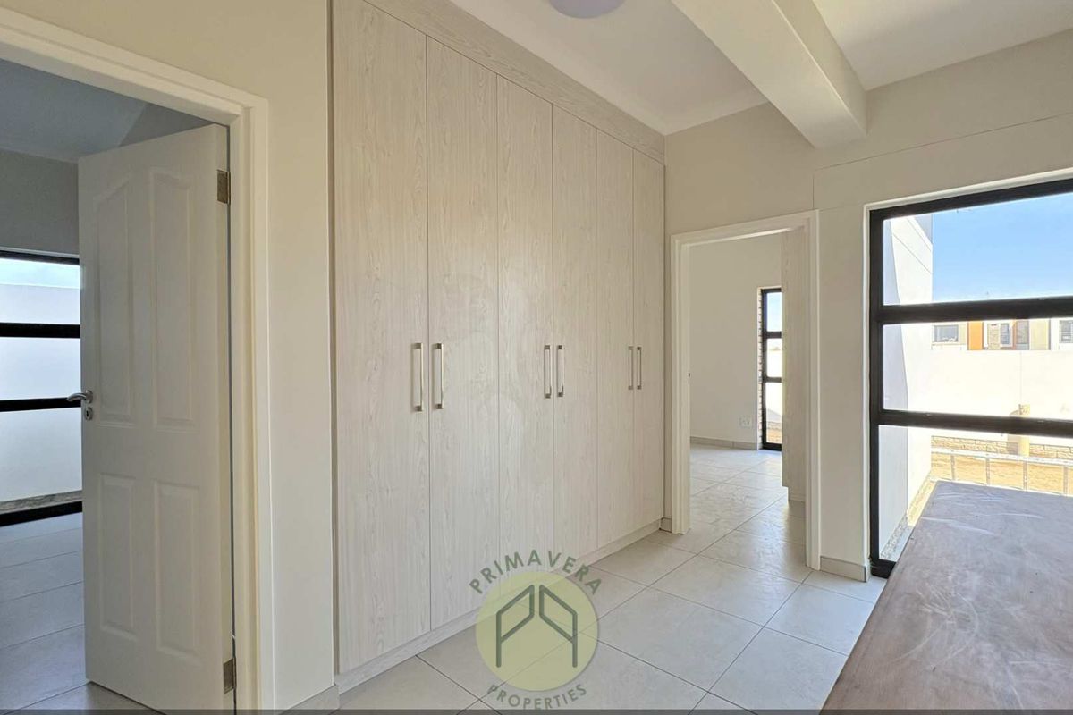 A bright, empty bedroom featuring modern built-in cupboards, neutral tiled flooring, and large aluminium-framed windows allowing for ample natural light.