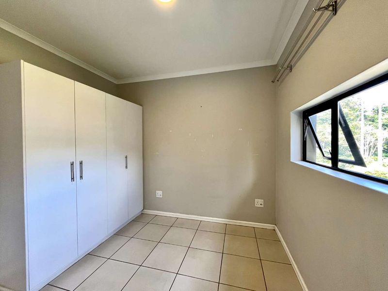 A bright, empty bedroom featuring neutral tiled flooring, built-in cupboards, and a large window with an aluminium frame.