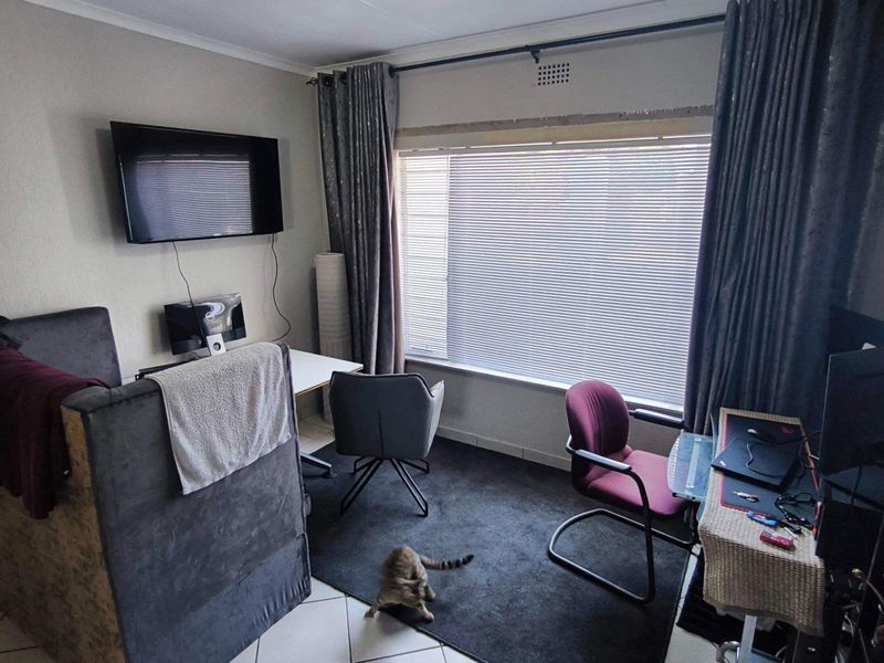 A functional living area featuring large windows with venetian blinds, curtain rails, and tiled flooring, currently utilized as a multi-purpose workspace.