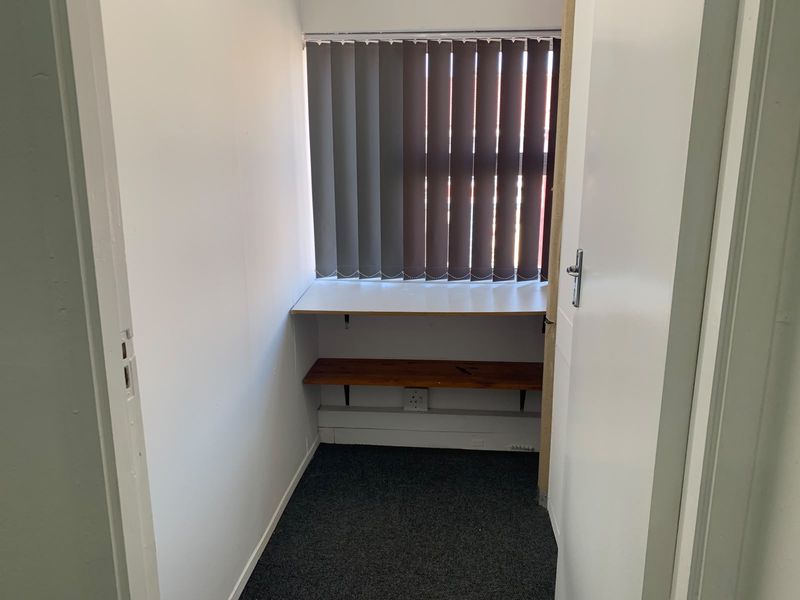 A compact office space featuring built-in desk and shelving, vertical blinds, and carpet flooring, suitable for a small workstation or storage area.