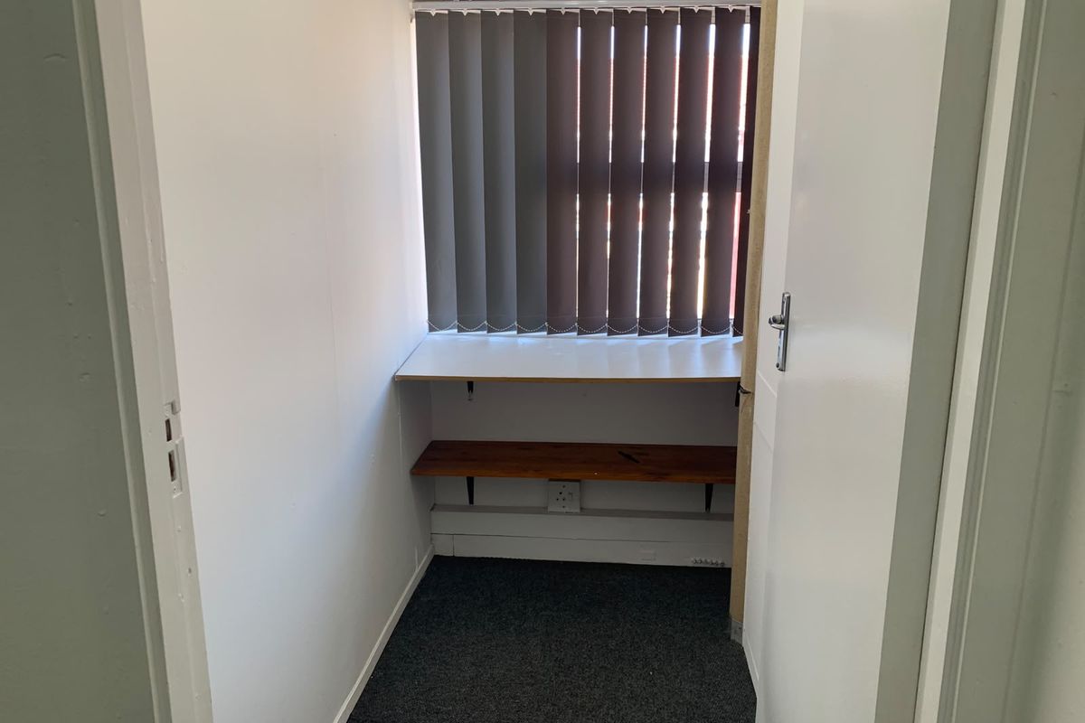 A compact office space featuring built-in desk and shelving, vertical blinds, and carpet flooring, suitable for a small workstation or storage area.