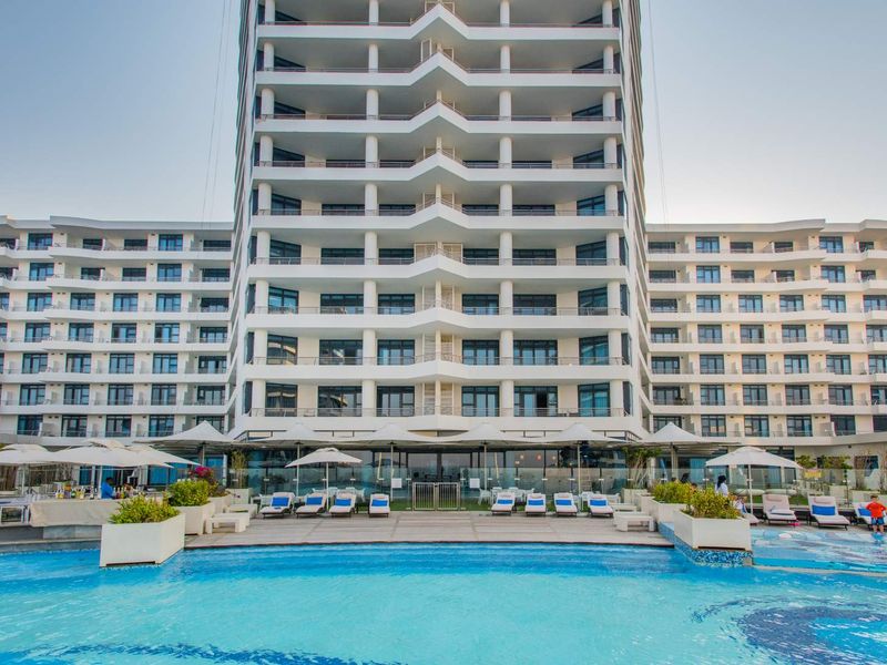 A modern high-rise apartment building featuring a large swimming pool area with paved decking and multiple levels of private balconies.
