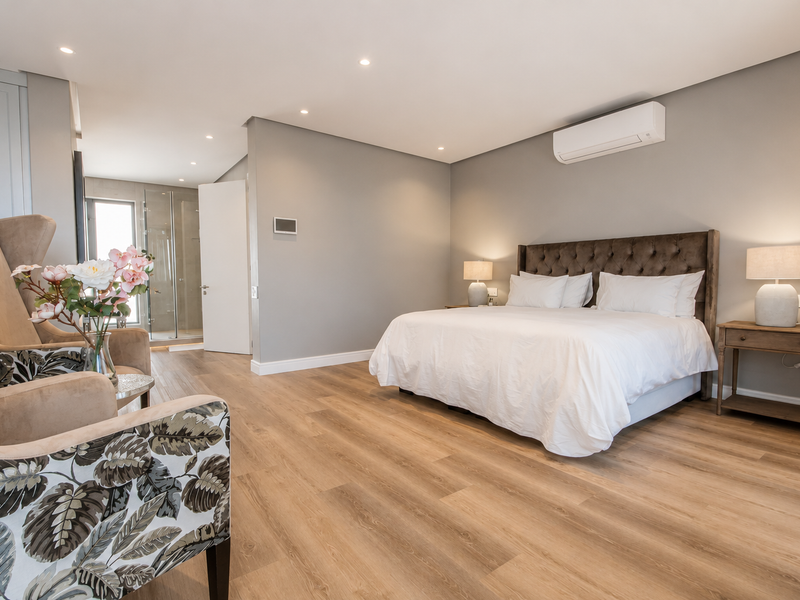 Spacious modern bedroom featuring light wood-look laminate flooring, recessed lighting, a wall-mounted air conditioning unit, and direct access to an en-suite bathroom.