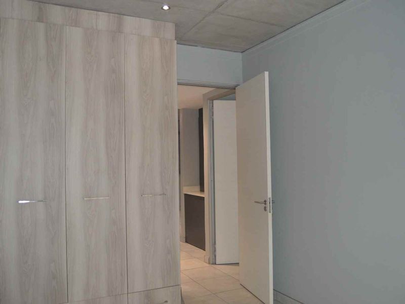 A clean, modern bedroom featuring light wood-finish built-in cupboards, an exposed concrete ceiling, and neutral tiled flooring, with a view into the adjacent bathroom.