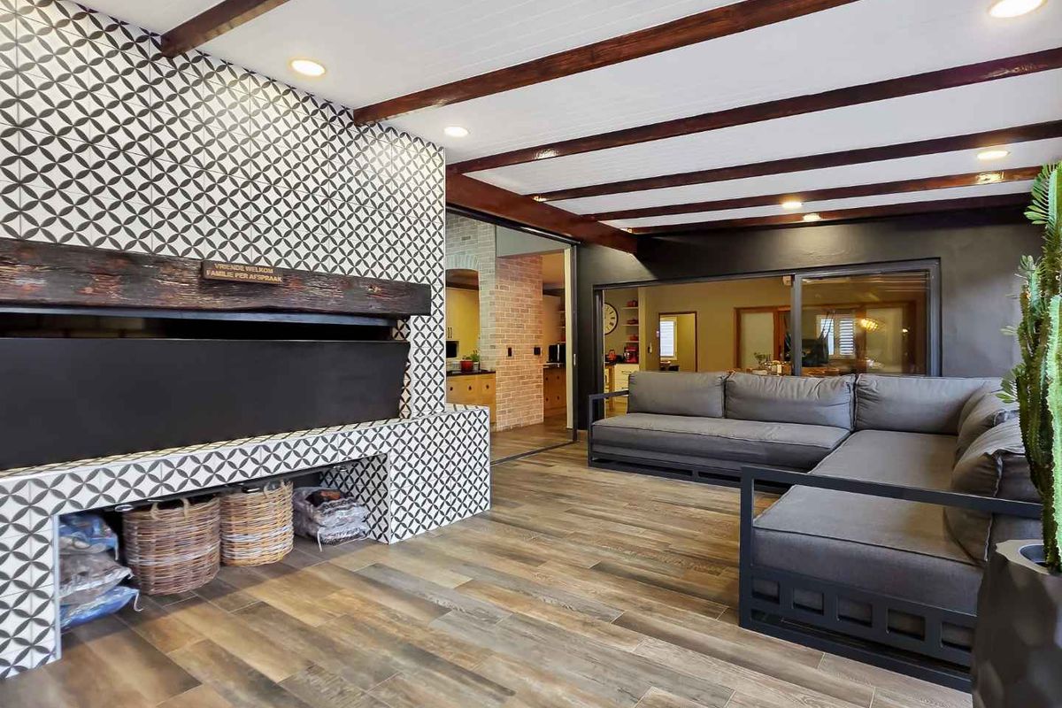 Spacious living area featuring a striking patterned tile fireplace, exposed timber ceiling beams, and wood-look flooring, with seamless flow to the adjacent kitchen.
