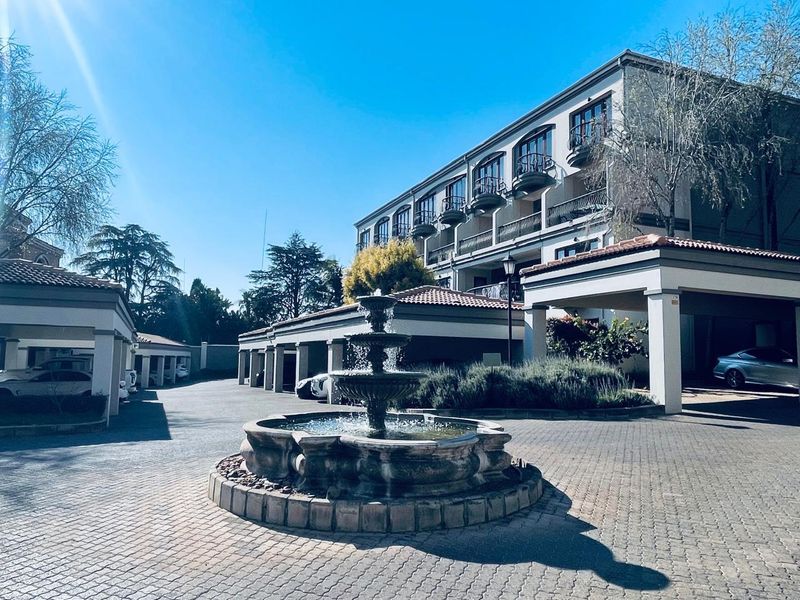 A well-maintained apartment complex entrance featuring a central stone fountain, paved driveway, and covered parking areas with balconies above.