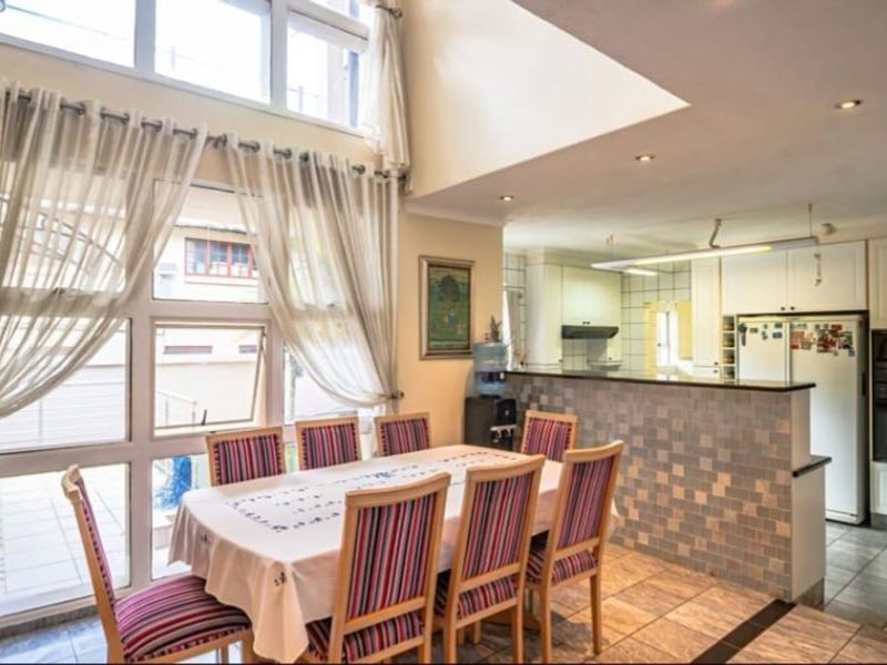 Spacious open-plan kitchen and dining area featuring double-height windows, tiled flooring, and a mosaic-tiled breakfast bar with integrated cabinetry and appliances.
