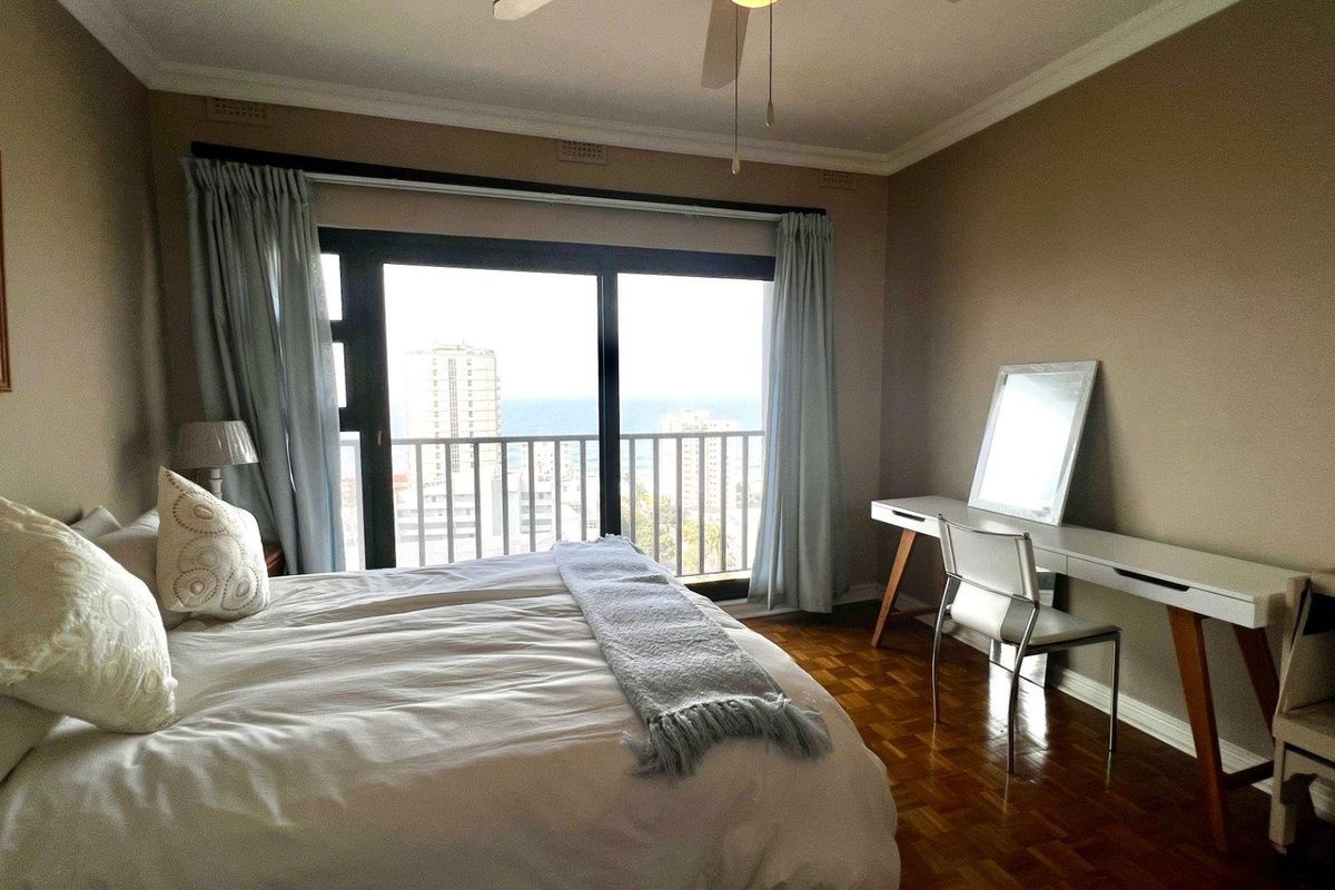 A well-lit bedroom featuring classic parquet flooring, a ceiling fan, and large sliding glass doors that open to a balcony with city and sea views.