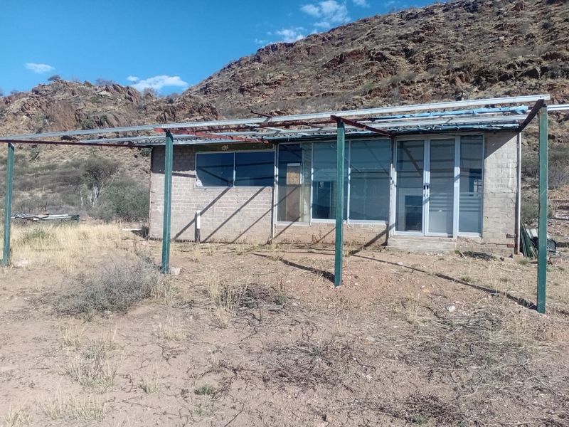 Rear view of a small structure featuring a metal pergola frame, sliding glass doors, and aluminum window frames set against a rocky hillside.